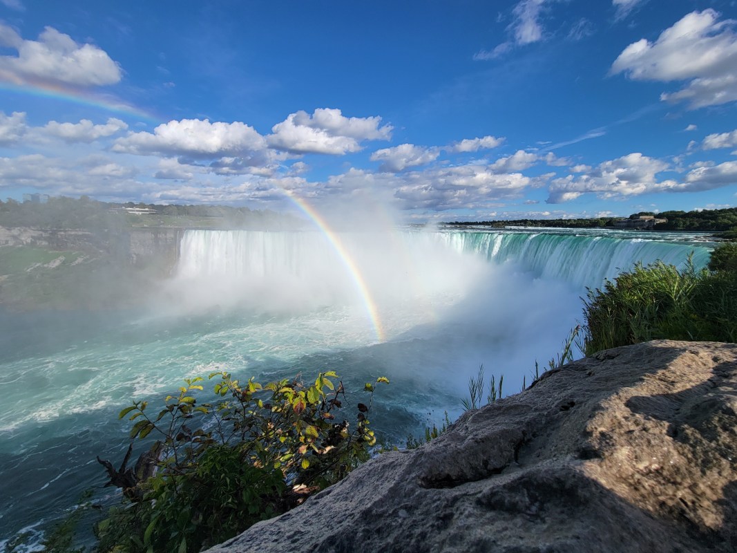 Water flowing over Horseshoe Falls cresting mist on a sunny day resulting in a colorful rainbow appearing to rise from the base.